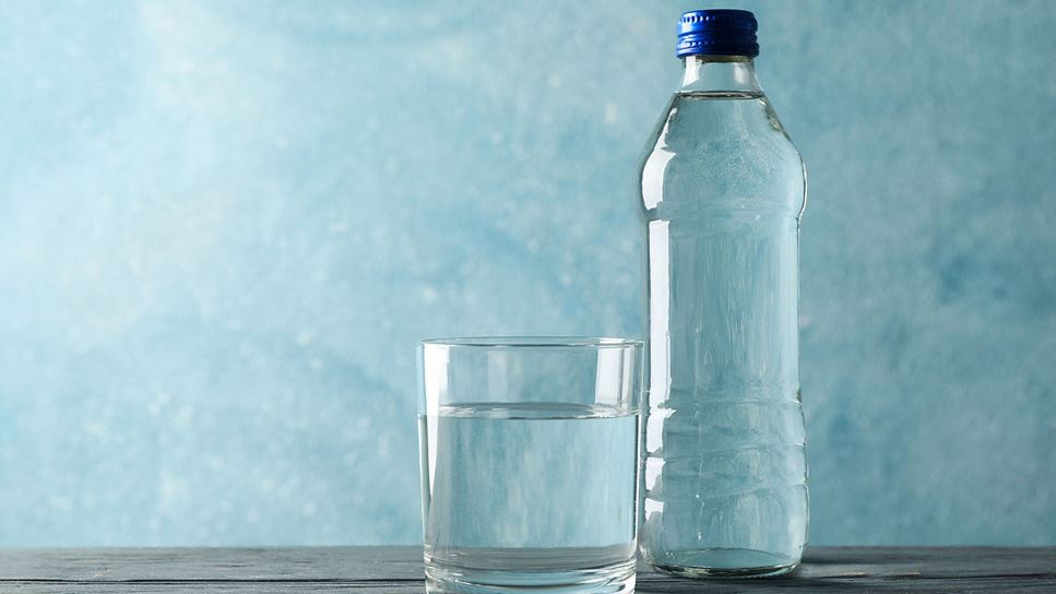 A clear glass of water on the left side of a clear bottle of water with a blue cap on a table with a blue background. 