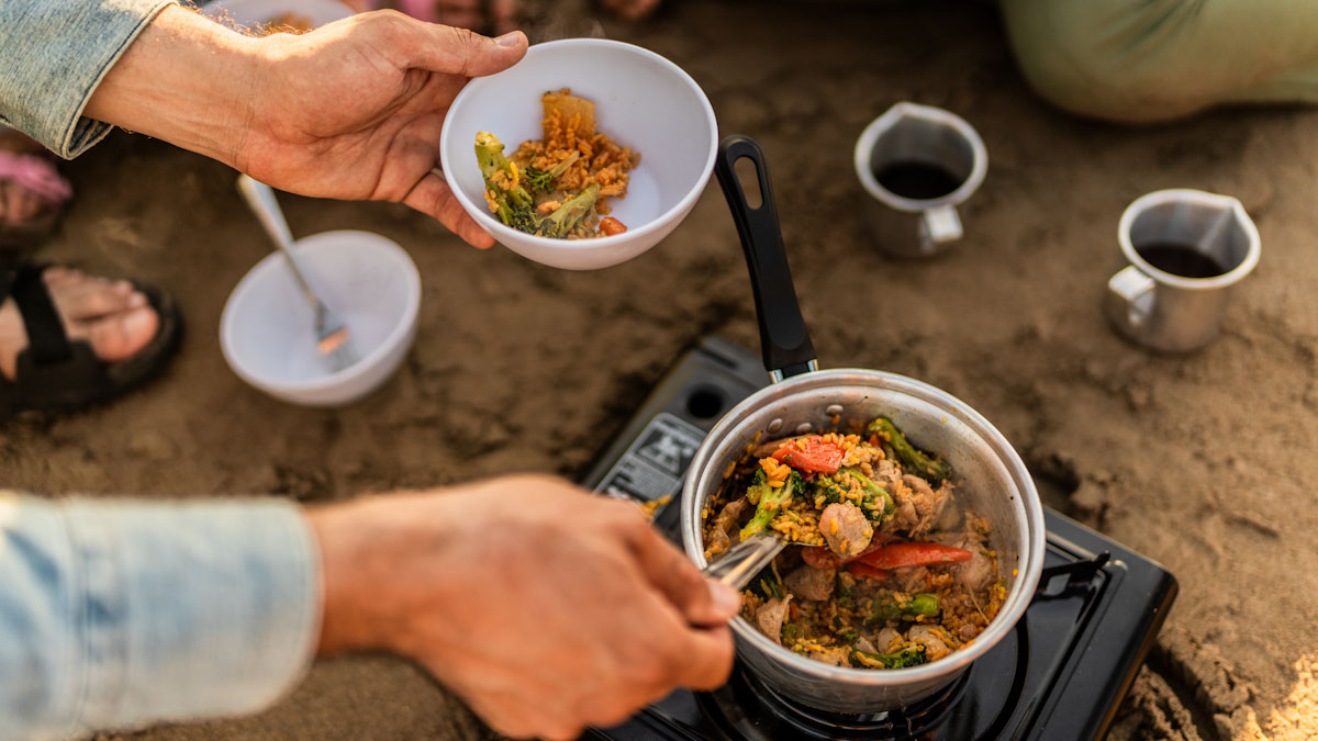 A person scoops a ladle of stew, cooked over a campfire.
