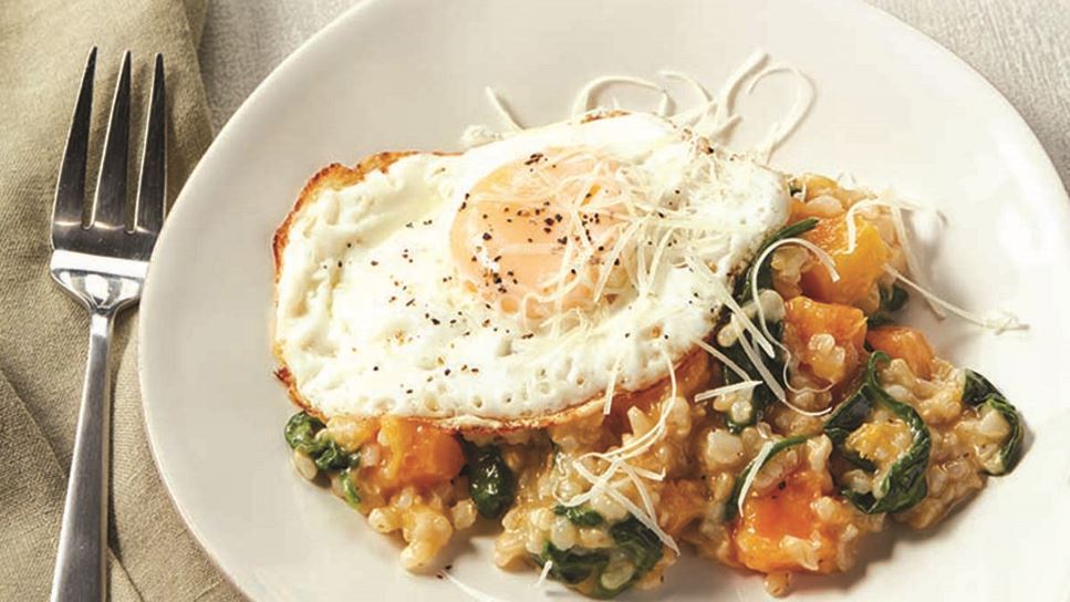 A white plate with brown rice risotto and veggies topped with a fried egg and grated Parmesan cheese. 