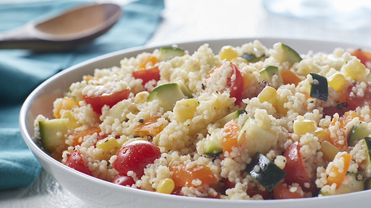 A colorful bowl of couscous mixed with brightly-colored vegetables on a table next to a blue napkin with a wooden spoon. 