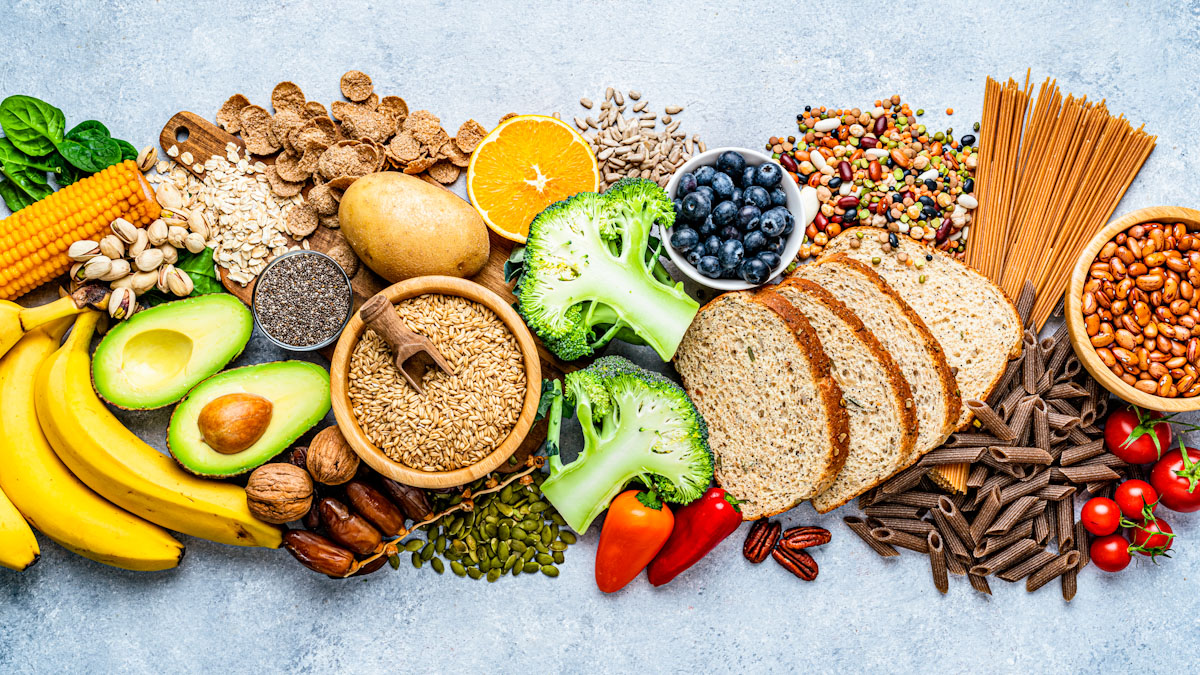 An assortment of foods providing fiber, including bananas, broccoli, avocado, corn, beans, whole grain bread, whole grain pasta, tomatoes, peppers, nuts and blueberries.