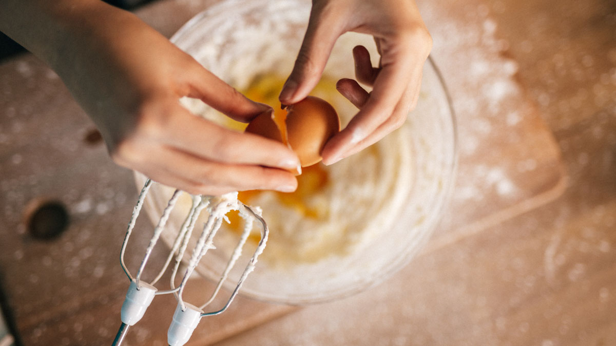 Close-up of hands breaking an egg over a batter. The beaters of an electric hand mixer are just above the bowl.