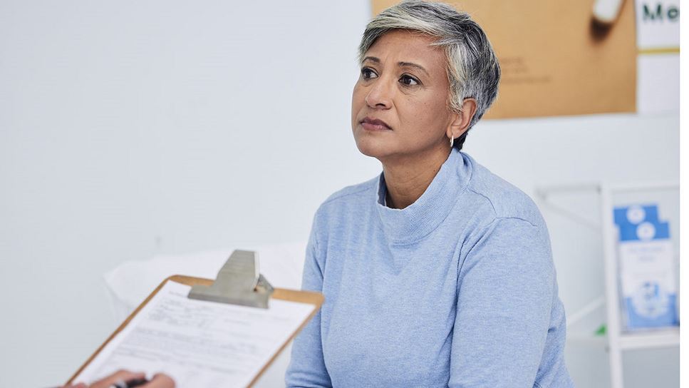 A woman with short hair dressed in a light blue sweater in a doctor's office looking at someone (not seen) taking notes on a clipboard.