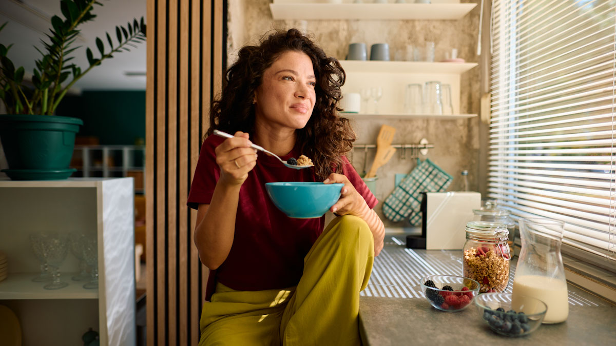 A woman sits in her kitchen, looking out the window, as she eats breakfast.