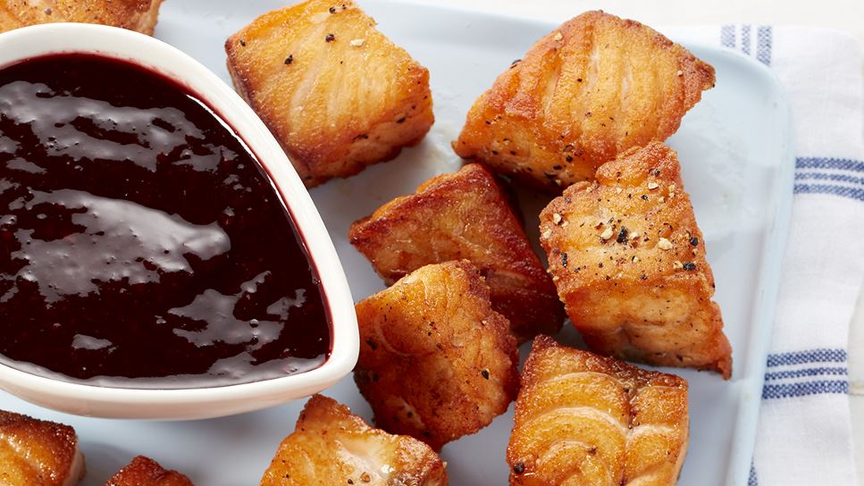 Salmon nuggets and blackberry dipping sauce on a white dish that's atop a blue and white placemat. 