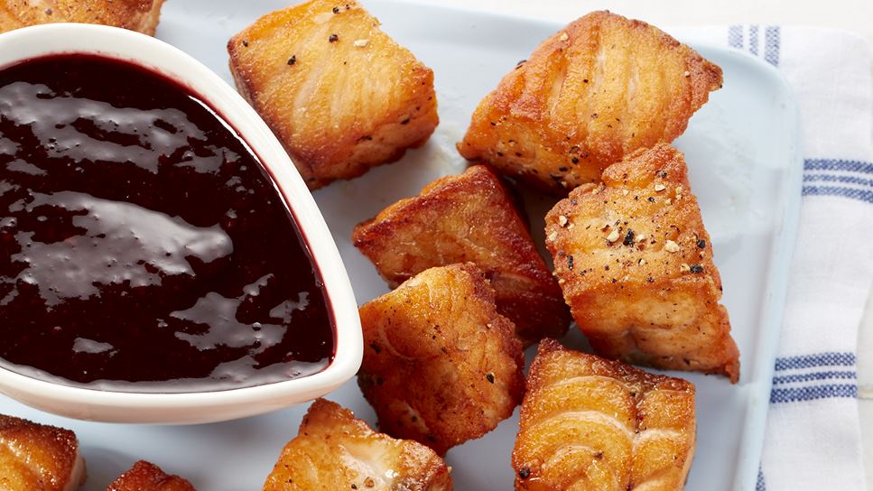 Salmon nuggets and blackberry dipping sauce on a white dish that's atop a blue and white placemat.