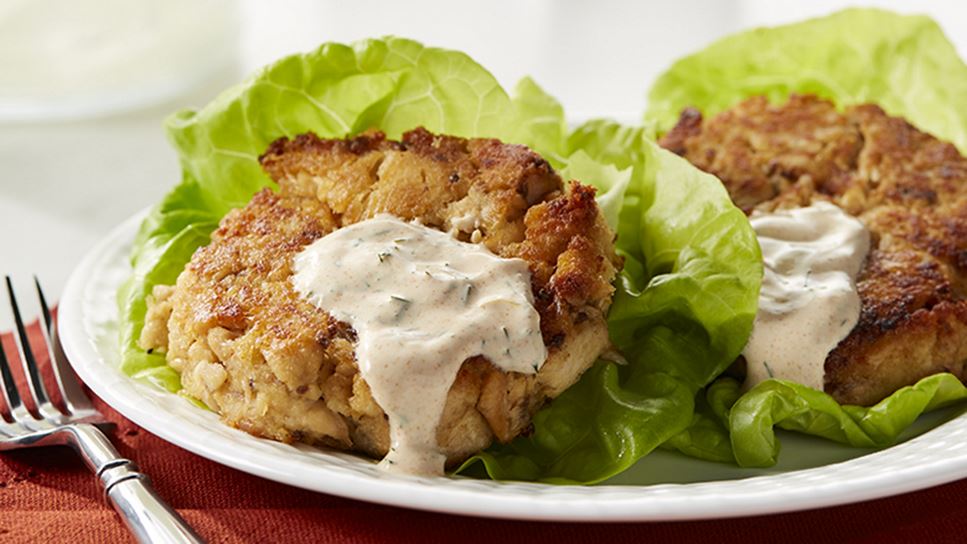 Two salmon cakes resting on lettuce leaves on a white plate sitting on an orange placemat with a fork resting on the left. 