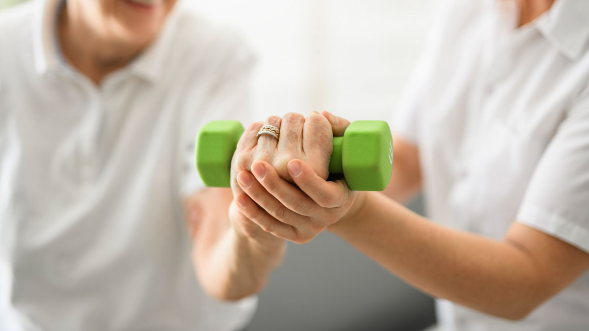 An older adult works on lifting a free weight to build muscle strength, with the assistance of a health care provider.