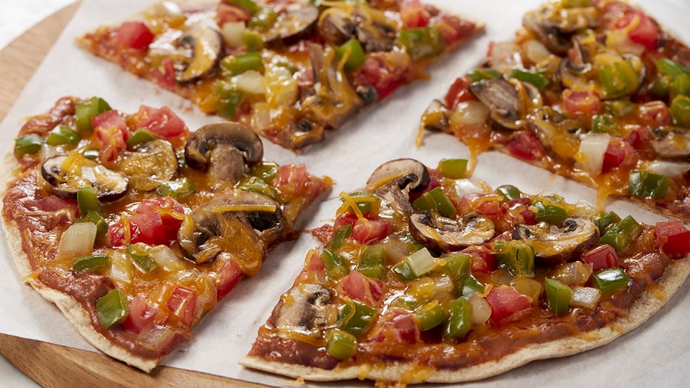 Four pieces of sliced pieces with mushrooms, peppers and tomatoes on a piece of paper that's on a round wooden slab. 