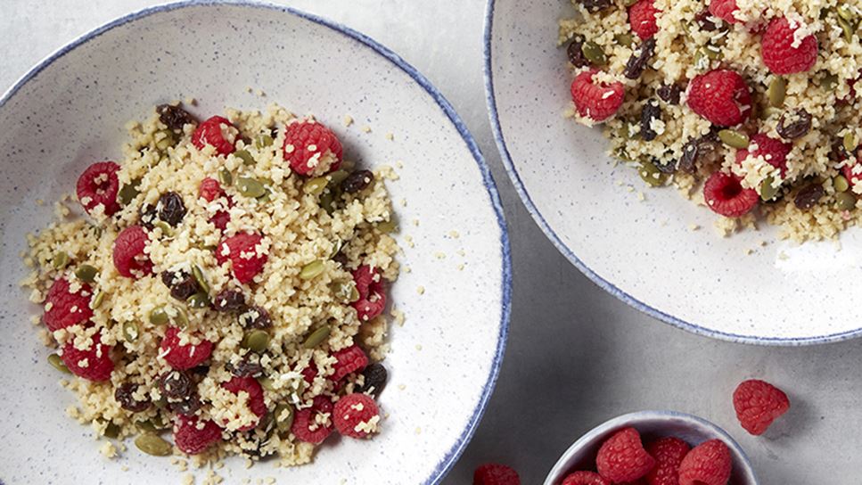 Two large bowls with a blue trim filled with couscous mixed with raspberries and pumpkin seeds. Also a small bowl of raspberries.