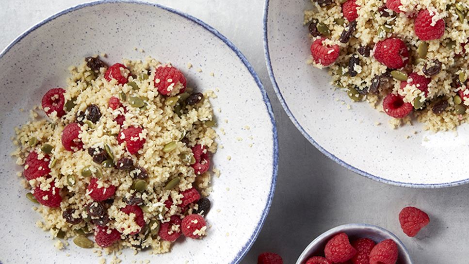 Two large bowls with a blue trim filled with couscous mixed with raspberries and pumpkin seeds. Also a small bowl of raspberries. 