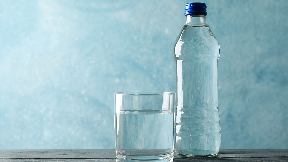 A clear glass of water on the left side of a clear bottle of water with a blue cap on a table with a blue background. 