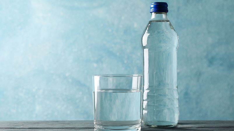 A clear glass of water on the left side of a clear bottle of water with a blue cap on a table with a blue background.