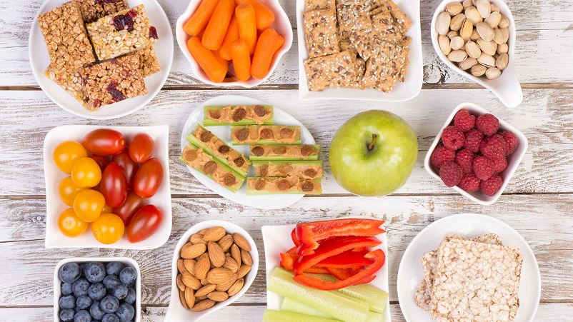 An assortment of healthful snacks for kids including homemade granola bars, fresh fruit and vegetables, nut butter and nuts.