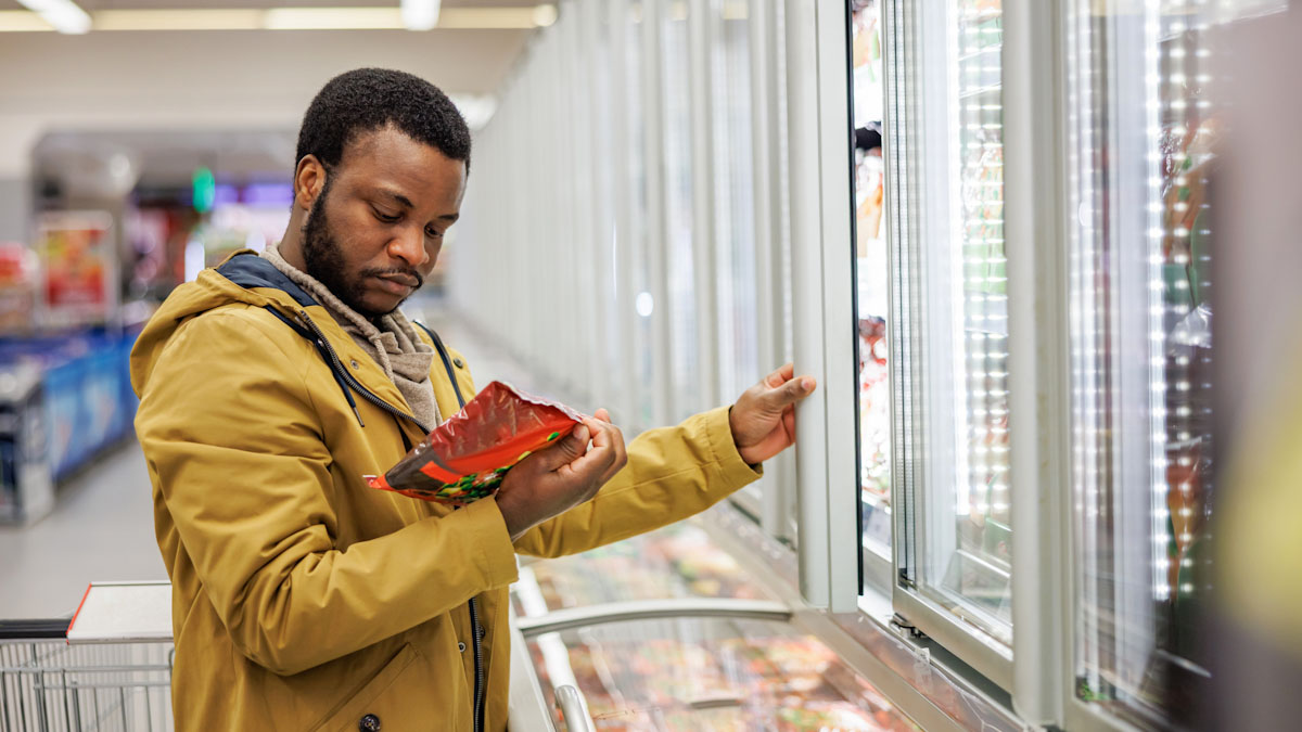 A man is looking at the nutrition fact label on a frozen food package while shopping at the grocery store.
