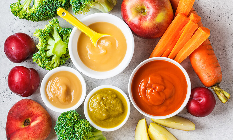 baby food in bowls with fresh fruit and vegetables arranged around