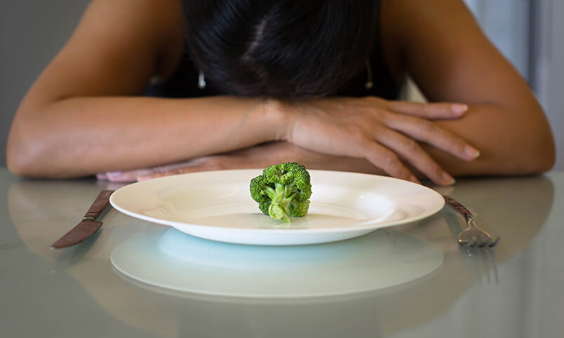 woman with head down on table in front of plate with one piece of broccoli