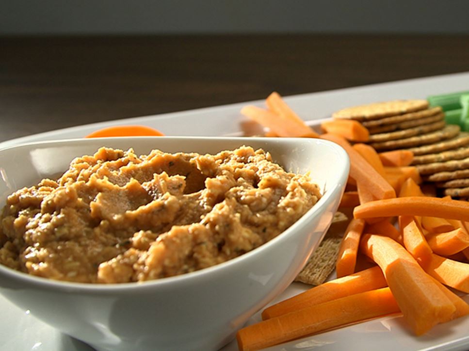 A white bowl of pizza hummus sitting on a plate with raw carrot sticks crackers and celery. 